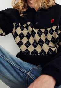 Polo noir avec motif losange beige, arborant un logo rouge, une fermeture à boutons et un tissu texturé. Porté avec un jean en denim.