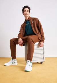 Veste de survêtement marron à fermeture éclair et pantalon assorti, portés sur un t-shirt vert. Baskets blanches avec des accents verts. Tabouret en métal avec une surface texturée.