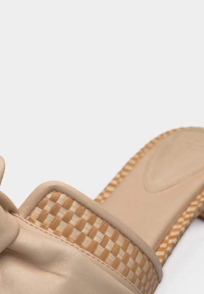 Κοντινή λήψη ενός μπεζ σανδάλου-σχάρας με πλεκτή λεπτομέρεια από άχυρο στο λουρί και την άκρη της σόλας, σε λευκό φόντο.