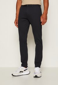 Sötétkék, slim fit nadrág rugalmas anyagból, lapos elejű, övhurkokkal és egy gombos záródással. Fehér sportcipővel viselve.