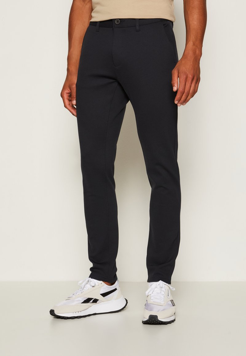 Sötétkék, slim fit nadrág rugalmas anyagból, lapos elejű, övhurkokkal és egy gombos záródással. Fehér sportcipővel viselve.