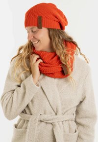 Oranje gebreide beanie met een slouchy ontwerp en een bruine label; bijpassende gestructureerde oranje gebreide sjaal; model draagt een lichtgekleurde, zachte badjas.