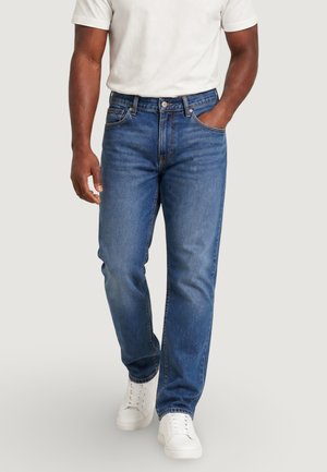 Homme portant un jean bleu moyen, des baskets blanches et un t-shirt blanc, marchant avec une main dans la poche devant un fond uni.