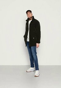 Casaco preto com colarinho alto, dois bolsos no peito e fecho com botão, combinado com uma t-shirt branca e jeans azuis, finalizado com sapatilhas brancas.