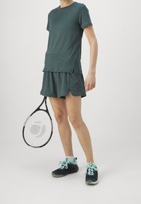 Sportovní outfit v tmavě tyrkysové barvě, skládající se z trička s krátkým rukávem a vrstvené sukně. Modelka drží tenisovou raketu a má na sobě černé tenisky.