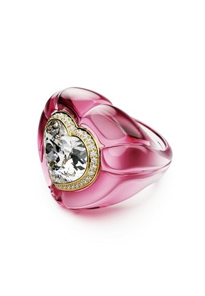 Różowy pierścionek w kształcie serca z żywicy, z dużym, sercowatym przezroczystym kryształem na środku, otoczonym złotą obręczą z akcentami diamentów.