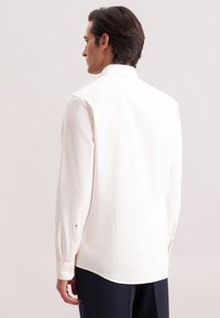 Man met donker haar, gekleed in een wit overhemd met lange mouwen en donkere broek, staat zijwaarts met zijn hoofd van de camera af gedraaid.