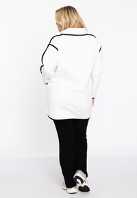 Witte lange mouwen top met zwarte piping, zachte textuur en kraag, gecombineerd met zwarte broek en sneakers. Van achteren getoond.