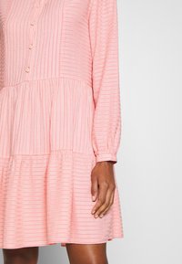 Vestito rosa con maniche lunghe, chiusura a bottoni, texture a righe e dettagli in gonna a strati. Tessuto morbido con una leggera lucentezza.