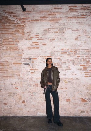 Giovane donna con capelli lunghi intrecciati che indossa una giacca di pelle marrone oversize, top nero corto, pantaloni neri a zampa d'elefante e stivaletti con plateau, appoggiata a un muro di mattoni.