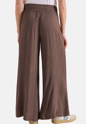 Kobieta w brązowych, szerokich teksturowanych spodniach i białych sneakersach z niebieskimi akcentami, stojąca na białym tle, widok od tyłu.