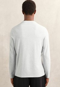 Svetlo šedé tričko s dlhým rukávom, vyrobené z mäkkej tkaniny, s okrúhlym výstrihom a rovnými okrajmi, oblečené s tmavými nohavicami.