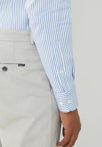 Chemise à manches longues rayée bleu et blanc avec poignets à boutons, associée à un pantalon gris clair avec une poche arrière et une fermeture à bouton.