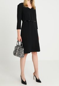 Svart knälång klänning med trekvartslånga ärmar, bälte i midjan, kombinerad med en handväska i ormskinn och svarta spetsiga klackar.