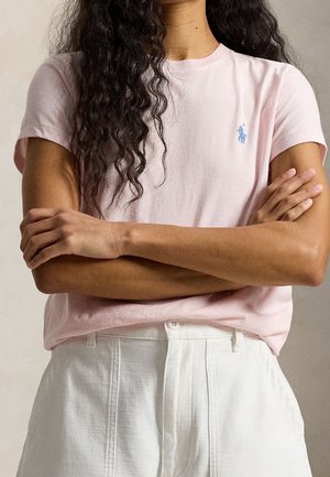 Person mit langen lockigen Haaren, die ein hellrosa T-Shirt mit kleinem blauem Logo und hoch taillierter weißer Hose trägt, Arme verschränkt.