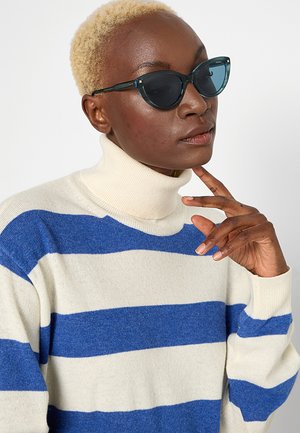 Persona con capelli biondi corti che indossa occhiali da sole a forma di occhio di gatto con lenti sfumate blu e un maglione bianco a collo alto con larghe strisce orizzontali blu.
