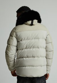 Pufferjakke i lys beige med en teksturert overdel og en svart pelskant på hetten, i quiltet design og med elastiske mansjetter.