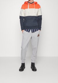 Felpa con cappuccio a righe orizzontali arancioni, crema e blu navy; abbinata a pantaloni della tuta grigi con tasche laterali e dettagli ricamati.