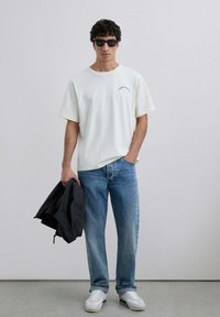 Camisa de algodón blanca con texto pequeño en azul, jeans de mezclilla azul claro, chaqueta negra en la mano y gafas de sol negras. Zapatos deportivos blancos.