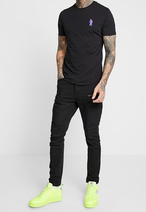 Uomo che indossa una maglietta nera aderente con piccolo logo viola, pantaloni cargo neri con tasca con cerniera e sneakers giallo neon, in piedi con le mani nelle tasche.