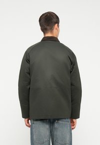 Donkergroene jas met een bruine corduroy kraag, gestructureerd ontwerp en raglanmouwen, gecombineerd met denim broek met grote achterzakken.