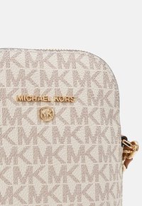 Bolso bandolera beige con una tela texturizada que presenta un patrón repetido de "MK". El herraje en tono dorado exhibe el nombre y el logo de la marca.
