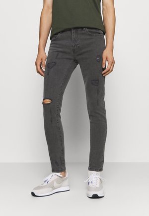 Mand iført sorte, slanke, slidte jeans med revner nær venstre knæ, kombineret med hvide sneakers og en mørkegrøn skjorte.