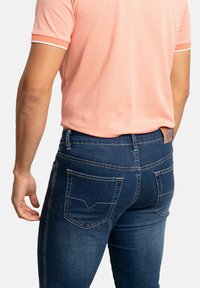 Jeans de mezclilla azul oscuro con costuras en contraste, dos bolsillos traseros con un diseño en forma de V, combinados con una camisa polo de textura rosa claro.