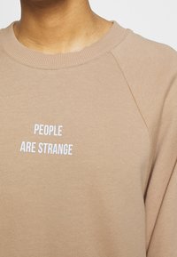 Sweat-shirt beige avec un col rond et des manches raglan. Le devant affiche un texte blanc indiquant "PEOPLE ARE STRANGE." Tissu doux et texturé.