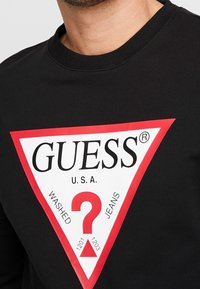 Felpa nera con un grande logo triangolare rosso e bianco. Il testo include "GUESS," "U.S.A.," "WASHED," e "JEANS" con un punto interrogativo.