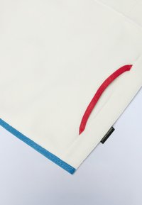 Coperta in pile bianca con accenti rossi e blu. Presenta una texture morbida, con una tasca rossa e dettagli del bordo blu. Etichetta nell'angolo.