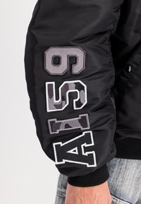 Melnā bumbvedēja jaka ar izolētu tekstūru. Iezīmētas lielas izšūtas ciparu kombinācijas pelēkā, baltā un kamuflāžas krāsā. Ribota aproce plaukstas daļā.