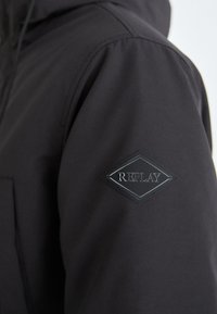 Giacca nera imbottita con una texture liscia e una toppa a forma di diamante etichettata "REPLAY" sulla manica sinistra.