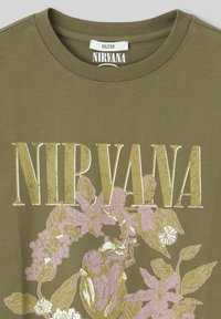 T-shirt en coton vert olive avec un texte "NIRVANA" en lettres dorées et des graphiques floraux roses, à col rond et à manches courtes.