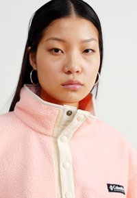 Veste en polaire rose clair avec un col haut, fermeture à boutons crème et un petit patch logo sur le côté gauche. Texture lisse et design décontracté.