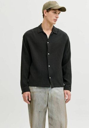 Young man wearing a textured black button-up shirt, light gray paint-splattered jeans, and an olive green baseball cap, looking to the side.