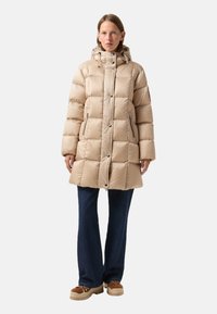 Beige Pufferjacke mit Kapuze, kastigem Design, gestepptem Material, Druckknopf- und Reißverschluss, seitlichen Taschen; kombiniert mit dunklen Jeans und klobigen Schuhen.