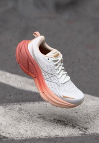 Baskets blanches avec semelle dégradée pêche, en l'air au-dessus de l'asphalte, avec une bande blanche, montrant le logo latéral et les lacets en détail net.