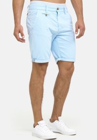 Lyseblå bomuldsshorts med opsmøgte kanter, forlommer og brune knapdetaljer. Sammen med hvide sneakers og en neutral baggrund.