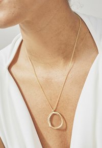 Gouden halsketting met een ronde hanger met textuurdetails, hangend aan een delicaat ketting, gedragen tegen een lichte stoffen top.