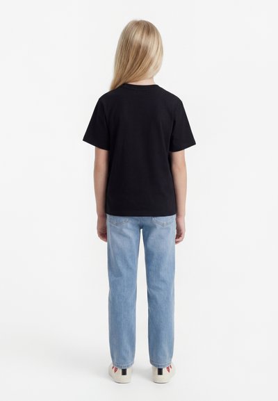 Enfant aux longs cheveux blonds portant un t-shirt noir, un jean bleu clair et des baskets blanches, debout de dos sur un fond blanc.