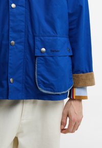 Μπλε μπουφάν Barbour με ασημένια κουμπιά τύπου snap, πατούρα τσέπης στο εμπρός μέρος, ταμπά μανσέτα, εμφανές χέρι και παντελόνι σε εκρού χρώμα.