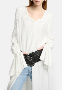 Weiße Bluse mit langen, gerafften Ärmeln, V-Ausschnitt und einem schwarzen Nieten-Gürtel. Hellblaue Jeans sind teilweise sichtbar. Glattes, fließendes Material.