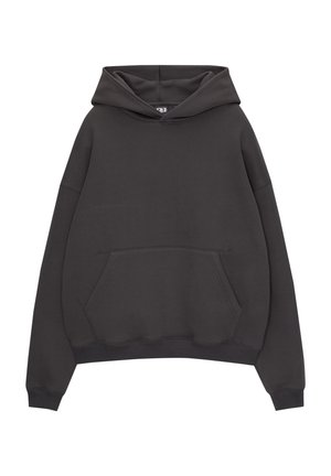 Person trägt einen locker sitzenden schwarzen Hoodie mit einer Fronttasche und dunkle Jeans mit weitem Bein, steht vor einem schlichten weißen Hintergrund.