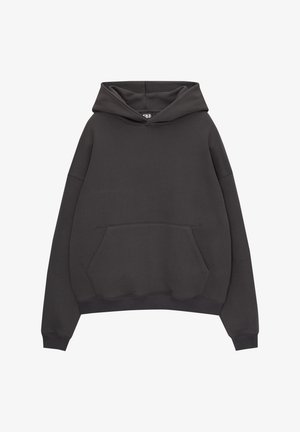 Person iført en løs sort hættetrøje med forlomme og mørke vidde ben jeans, stående mod en ensfarvet hvid baggrund.
