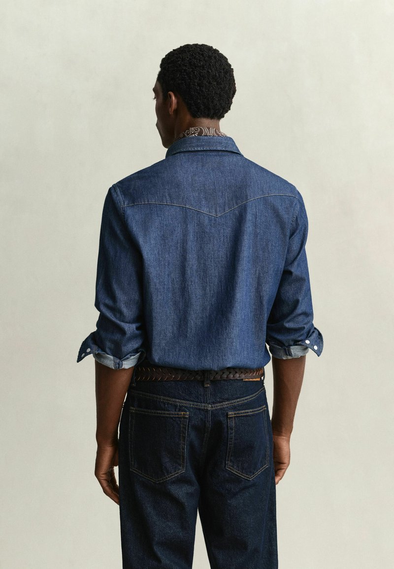 Man standing facing away wearing a rolled-up long sleeve blue denim shirt, dark blue jeans, and a braided brown belt.