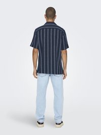 Chemise à manches courtes bleu marine avec des rayures verticales blanches, en tissu doux, coupe droite. Portée avec un jean bleu clair et des chaussures foncées.