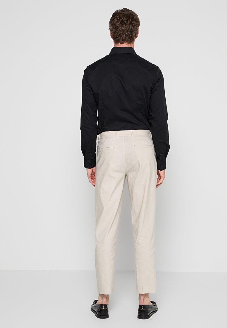 Mann mit langärmligem schwarzen Shirt, beigen Hosen und schwarzen Slippers, der vor einer hellgrauen Wand steht.