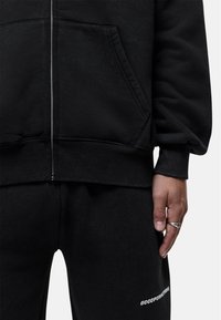 Felpa nera con zip e pantaloni della tuta in morbido tessuto. Design con cappuccio, polsini a costine e dettagli con logo sui pantaloni. Zip argentata e anello in mano.
