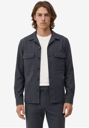 Junger Mann mit schulterlangem braunem Haar, der eine dunkelgraue, geknöpfte Jacke mit Klappentaschen und passende Hosen über einem weißen Hemd trägt.